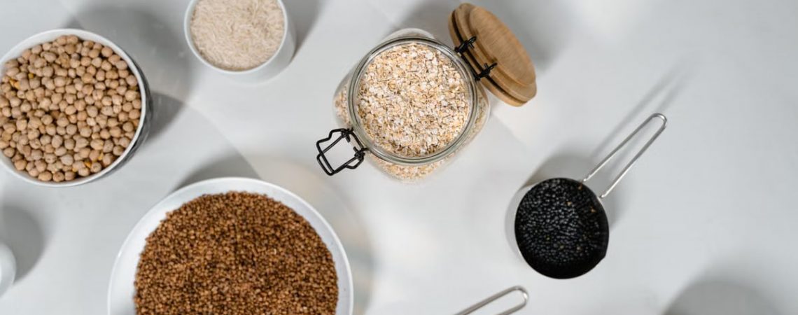 Top view of various grains and seeds in bowls and jars on a white surface.